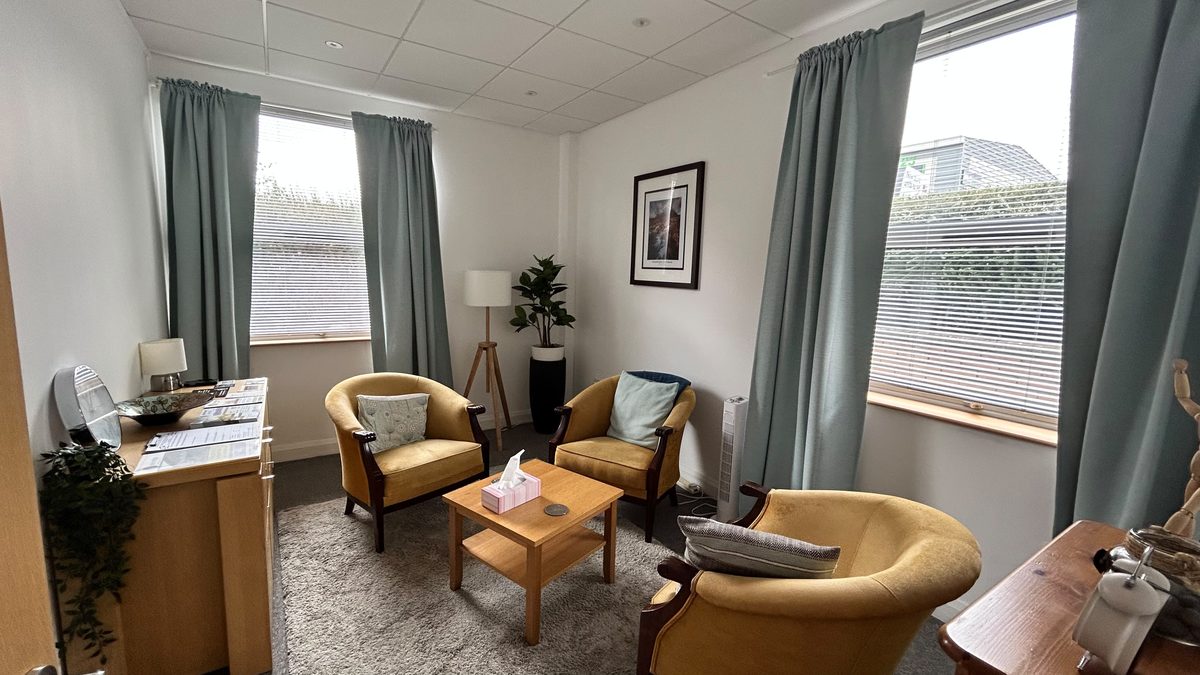 The counselling room: two mustard-yellow armchairs and a low wooden table on a soft grey rug, sage-green curtains at two windows, a floor lamp and a potted plant in the corner.